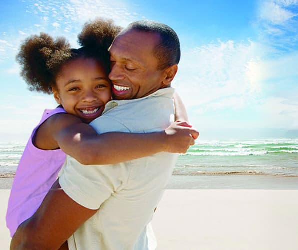 Man and girl embrace and smile on sunny beach with surf behind them.